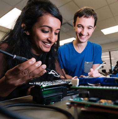 a woman working on a electronic component while a man behind her watches